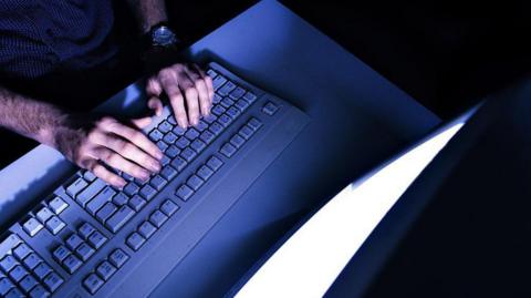 The hands of a man typing on a keyboard in front of an illuminated screen, in a darkened room