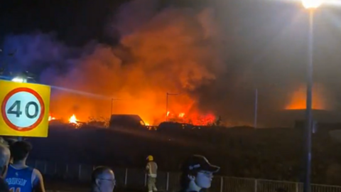 A grass fire is seen in the distance as people watch by a street.