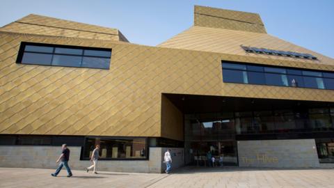 The exterior of The Hive in Worcester, a distinctive gold building with a honeycomb-type pattern on the walls. A sign outside reads "The Hive". People are walking along the pedestrianised area outside.