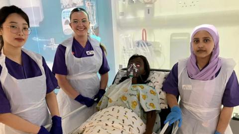 Three student nurses in scrubs smiles next to a manikin doll wearing an oxygen mask with brown hair leans back against a pillow