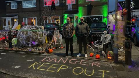 Andy Whittle and his son, Toby, stand outside their semi-detached home in the dark. The path in front of the house says 'danger keep out' and their front garden is full of lit pumpkins, fake skeletons, cobwebs