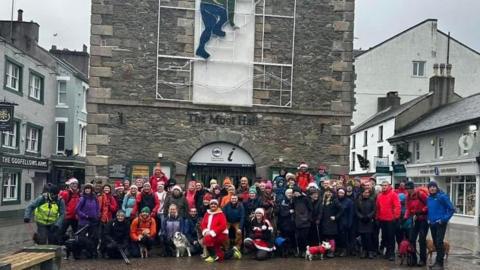 Lots of people are in front of a tower in Keswick. Most are dressed in walking gear but one man is dressed as Father Christmas 