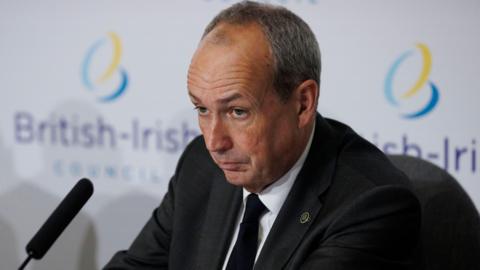 Deputy Lyndon Farnham, Chief Minister of Jersey, sits during a press conference at a British-Irish Council summit. He is wearing a suit with a black tie. A board behind him has two British-Irish Council logos on it. A microphone is in front of him.