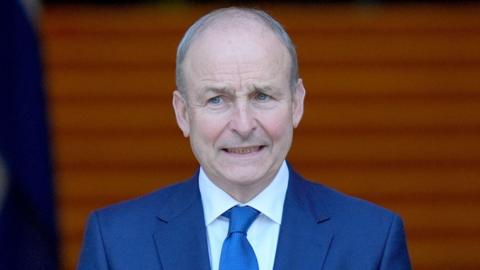 Taoiseach Micheál Martin, pictured from the chest up. He has balding short grey hair and he is wearing a dark blue suit, blue tie and white shirt.