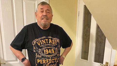 A man with grey hair and a beard stands smiling at the camera, in the hallway of a house. He is wearing a T-shirt which says "vintage 1943, perfectly aged".