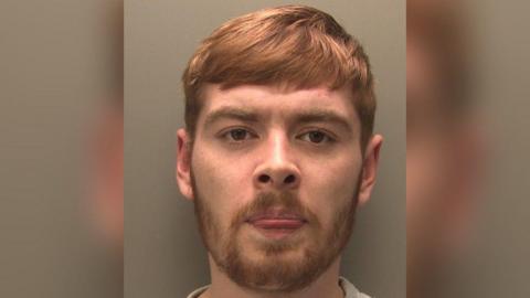 A man with short ginger hair that is swept across his forehead in a police headshot. He is frowning and his tongue is slightly out of his mouth. He had a short moustache and beard.