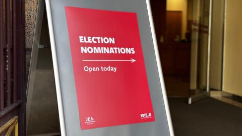 A red sign, with white writing saying 'election nominations open today' alongside an arrow pointing to the right. It placed outside a building.