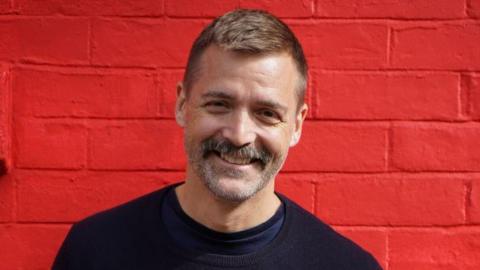 Patrick Grant is smiling at the camera. He has a moustache and is wearing a black sweatshirt over another dark top. He is standing in front of a bright red brick wall.