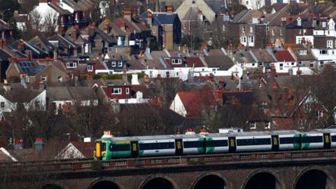 A train crosses the London Road Viaduct as it travels through Brighton. It is a southern train with green circles on its side and yellow panels. The arches of the viaduct can just be seen at the bottom of the image and there are houses in the background.