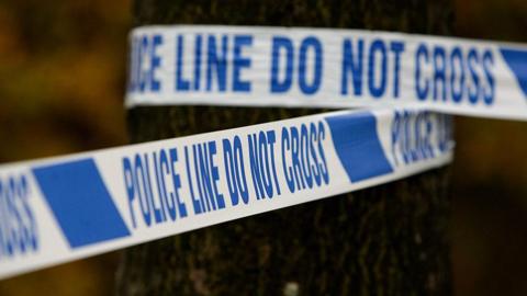 Generic close-up image of police tape wrapped around a tree trunk saying 'police line do not cross' in blue writing on a white background