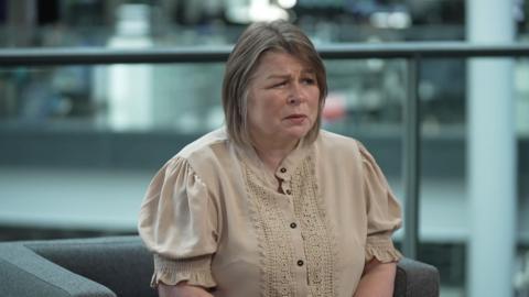 A woman in a cream top and shoulder length hair is looking away from the camera. She is sat on a grey chair and there is a divider made of glass in the background.