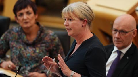Shona Robison speaking in the Scottish Parliament. She has blonde hair and is wearing a black top, and is holding her hands out, holding a pen, while talking. Angela Constance is seated to her left and John Swinney is sitting to her right.