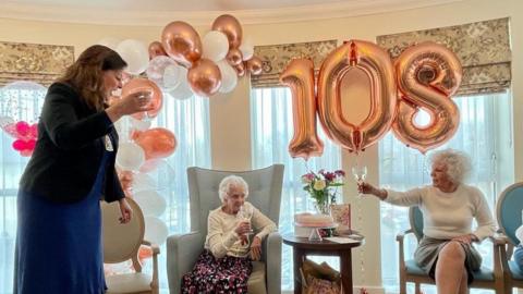 Bromley Mayor Councillor Hannah Gray, Rosina Clifton, and a family member in front of rose gold balloons spelling out the number 108.