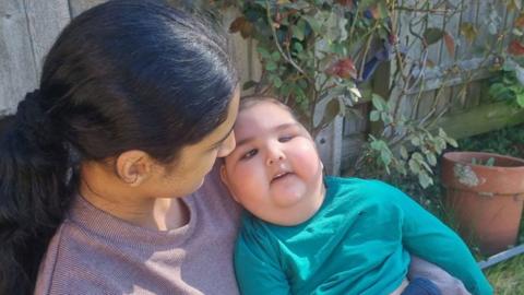 Zak being held by his 15-year-old sister, Ruwaida. Ruwaida is on the left and her head is turned towards her brother so very little of her face can be seen. She has long black hair in a pony tail and is wearing a mauve top. Zac is looking up towards the camera. He has a very swollen face and is wearing a teal top. They are in a garden and grass, a pot, a rose bush and a fence can just be glimpsed. 