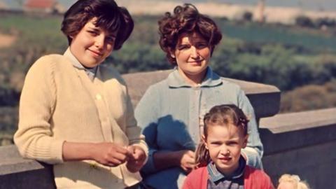 A young woman with short dark hair wearing a cream cardigan leans against a wall smiling at the camera. Another young woman with short dark hair stands next to her wearing a blue cardigan with a six year old girl standing in front of her wearing a blue collared dress with pink cardigan and holding a blonde haired doll.