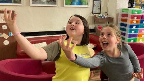 Two girls stand with their mouths open and their right arms stretched out in front of them as if holding an object. One has brown hair and is wearing a yellow and brown Brownies t-shirt. The other has blonde hair and a grey long-sleeved top.