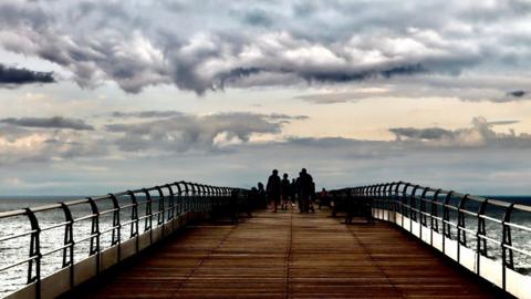 A cloudy scene over the North Sea with people standing on a pier.