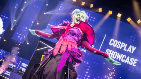 A person in an elaborate cosplay outfit pulls an extravagant pose on-stage. They're wearing a pink/purple dress, their hair is dyed blue and their skin is made up to look pale green/alien.