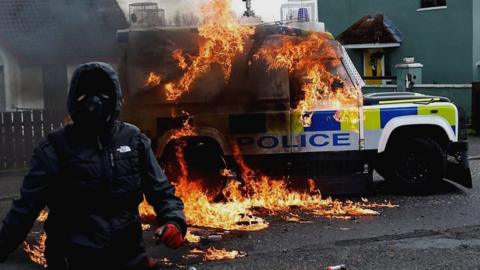 Masked man stands in front of burning police van