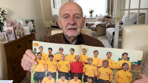 A man in his seventies holds a poster of the 1968 Watford football team wearing yellow shirts. He is pictured in the team photo. 