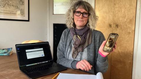 A woman sits in her kitchen looking glum, with a laptop and a mobile phone, to demonstrate how she has no internet 