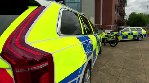 The picture shows two police cars and a motorbike which are covered in luminous yellow and blue graphics. They are parked on stone paving in front of a large modern building.