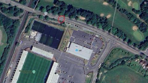 An aerial view image of Shrewsbury Town football ground and surrounding pitches, buildings and fields, taken from Google. Towards the top of the image is Oteley Road with the location of the new crossing marked with a red circle.