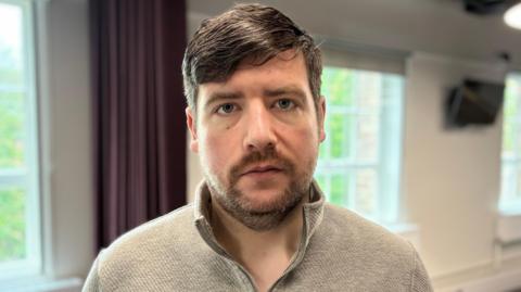 Alan Irwin had a beige half zip jumper on, He has a brown moustache and beard, and his hair is swept over to one side.