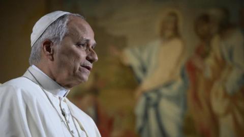 Pope Leo in the Vatican, dressed in papal robes. In front of a big TV showing Americans.