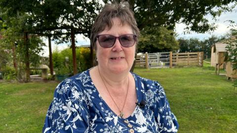 A head and shoulders shot of Trudie with brown hair wearing glasses, a blue and white top and necklace. She is standing on a lawn with trees, animals and their shelters in the background.