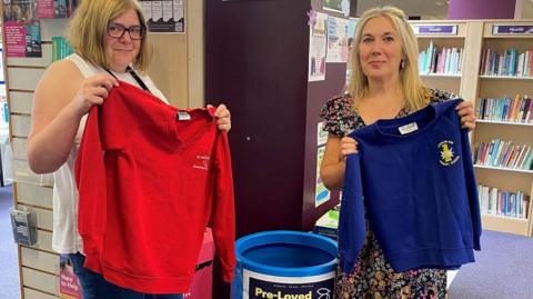 Liz Beddows and Lisa Bentley at Cannock Library holding up two branded school tops. Liz Beddows hold a red top and Lis Bentley holds a blue top. They are beside a bucket for school donations and behind them are book shelves. 