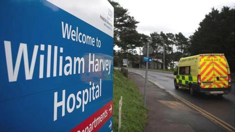 A shot of a sign outside William Harvey Hospital in Ashford with an ambulance in the background.