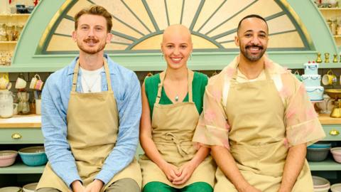 Tom in a blue shirt and beige apron, Jasmine in a green jumsuit and beige apron and Aaron in a flowery shirt and beige apron sitting in the Bake Off tent.