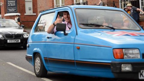 Harry Atkinson driving the powder blue Reliant Robin, smiling and giving a thumbs up out of the side window.