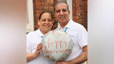 Gurpal and Kinder Matharu stand lose together in white NHS volunteer shirts. they are holding a balloon that reads "happy anniversary"