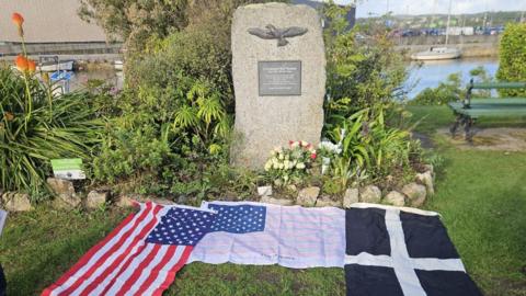 The American flag with red stripes and white stars on a blue background is lying on the grass near the Cornish flag with a white cross on a black background. They are in front of a stone monument which has a metal image of an eagle on it.
A sailing boat can be seen on the water in the background.