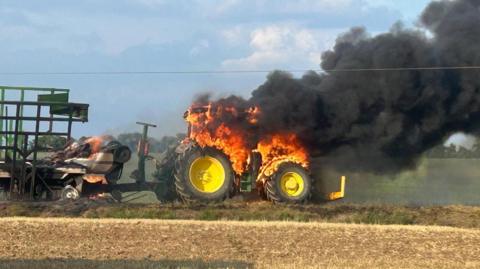 The body of a tractor is engulfed in flames, only the wheels are not burning. Coming from the top of the red and orange flame is a large billowing cloud of thick black smoke. The tractor is pulling farming machinery, which has also caught fire. It is burning in a large field.