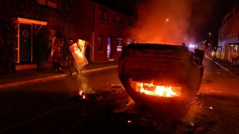 A upturned car is set on fire in the middle of the street. It is dark outside. Surrounding the car are police officers holding up large, plastic riot shields.