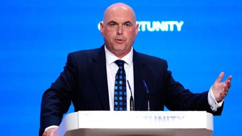Paul Davies standing at a lectern in a navy suit, white shirt and navy tie with blue dots. He has a shaved head and is gesticulating as he talks during a conference. The backdrop is blue and the word opportunity is written on the lectern and projected onto the wall behind him.