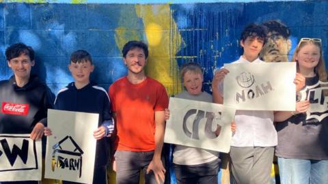 A group of children hold up pieces of paper with cut-outs of their graffiti tags. They are standing in front of a brick wall spray-painted blue and yellow.
