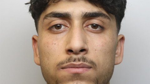 A mugshot of Hadi Al Koumi, showing his face from his chin to his hairline. He has a a dark skin tone, and is looking directly at the camera with a straight face. He has dark brown hair and eyebrows, and brown eyes. He has a moustache and some facial hair on his chin and cheeks.