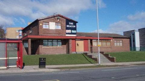 A red brick two-story building with a red bus stop outside. On the building a black and white sign reads: The Beacon