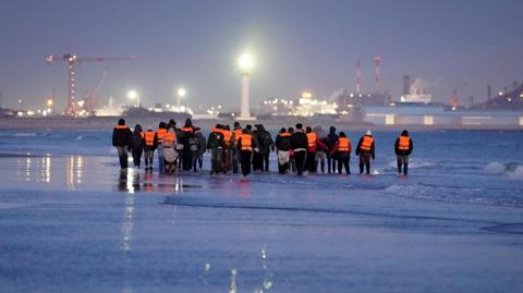 A group of around 20 people wearing lifejackets walking along the water's edge.