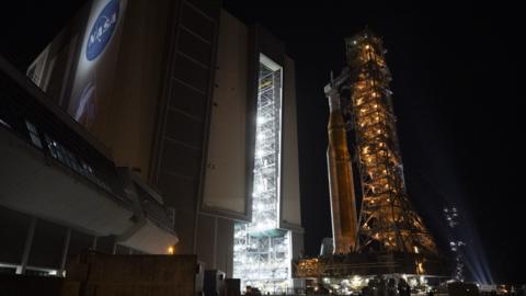 A large rocket, secured to a mobile launcher, getting rolled out of a Nasa hangar whilst it's dark outside.