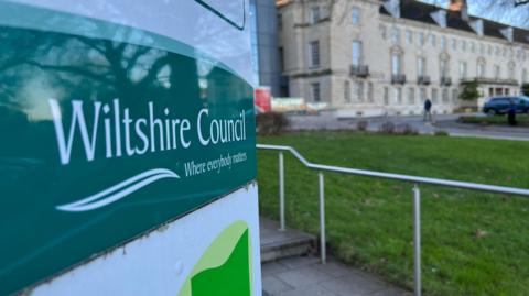 A close-up of a Wiltshire Council sign outside its headquarters, which is blurred in the background, a large grand building with bay windows.
The sign shows the green logo with white lettering.