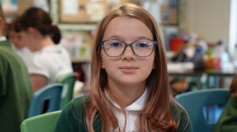 A school girl with long brown hair and blue rimmed glasses looks into the camera 