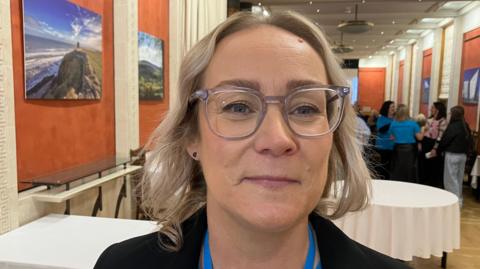 Caroline Campbell - a woman with short, blonde hair wearing grey, square-rimmed glasses. She is smiling at the camera and is standing in the middle of a hall. In the background are some tables with white tablecloths and people talking in the background.