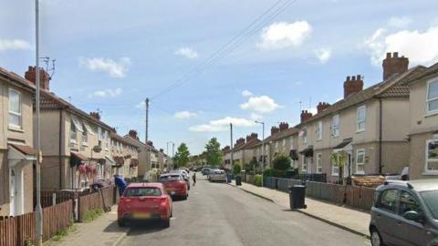 There are rows of terraced houses on either side of Fairbrook Drive in Birkenhead North. There are are number of cars parked on the residential street. 