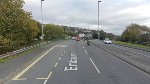 A picture of a large road in Plymouth. It is a grey road with markings on it, with vehicles travelling along it. Trees, lampposts and barriers can be seen either side of the road. In the distance there is trees and a bridge.