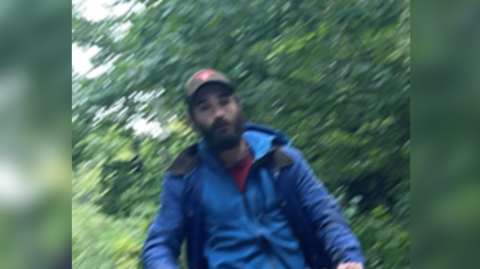 A blurry image of a man in a blue anorak with a red t-shirt underneath. He is white with a black beard, heavy black eyebrows and a brown baseball cap with a red logo on it.  